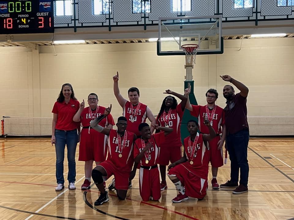 7 basketball players, a coach, and a sponsor hold up a number 1 finger and smile to camera.